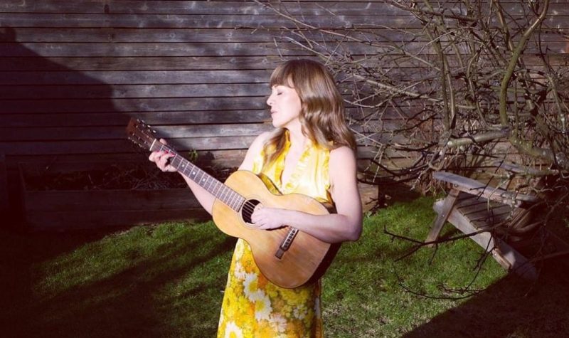 Une femme blonde habillée en robe jaune avec des fleurs. Debout dans une pelouse, elle tient la guitare entre ses mains.