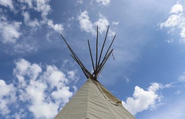 The top of a tipi, shown with sticks sticking out of the top, stands tall with a partly cloudy blue sky in the background.