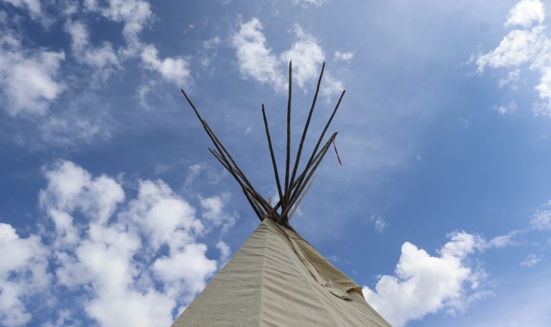 The top of a tipi, shown with sticks sticking out of the top, stands tall with a partly cloudy blue sky in the background.