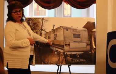 A woman wearing a white sweater points at a picture of a run down trailer that has mold forming on the outside of it. She's showing this because some residents of Conklin have to live in these conditions over lack of proper housing. Photo was taken inside.