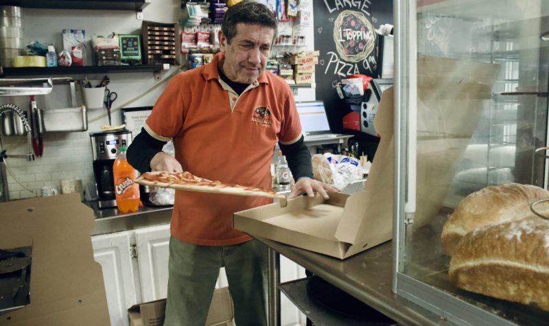 A man wearing a Triple A shirt putting a pizza in to a box. He is smiling. He is in a convenience store and there are grocery items behind him.