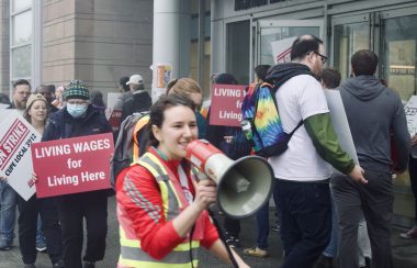 A woman on a speaker, she is walking and chanting. There are students in the back with signs calling for better wages. They are in front of a building on Dalhousie campus.