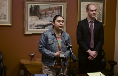 A woman standing in front of two microphones. She is speaking in to the microphone. Next to her is a man in a suit, her lawyer seen smiling to the press. This press release session took place in one of the meeting rooms at his firm.