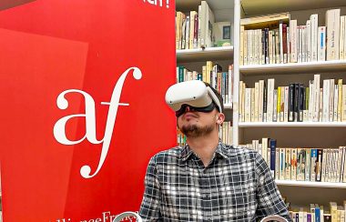 Un homme porte un casque de réalité virtuelle, il est dans une bibliothèque devant le logo de l'alliance française.