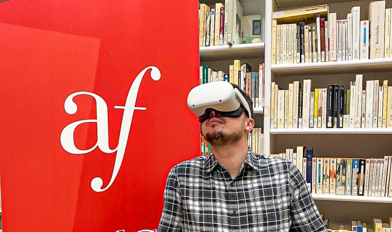 Un homme porte un casque de réalité virtuelle, il est dans une bibliothèque devant le logo de l'alliance française.
