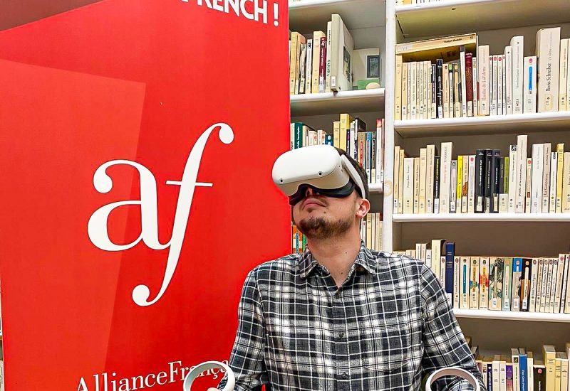 Un homme porte un casque de réalité virtuelle, il est dans une bibliothèque devant le logo de l'alliance française.