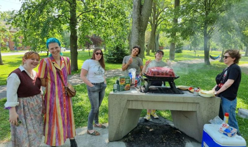 six volunteers stand at a barbeque with the grass and trees of Victoria Park in the backdrop