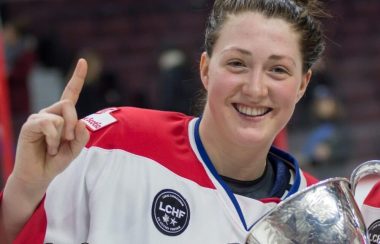 Une joueuse de hockey en uniforme blanc et rouge, tout sourire tenant une coupe de championnat