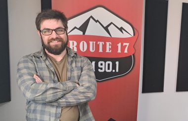 Homme portant une chemise et une barbe les bras croisés et souriant pour une photo devant inscrit Route 17 en blanc, rouge et noir