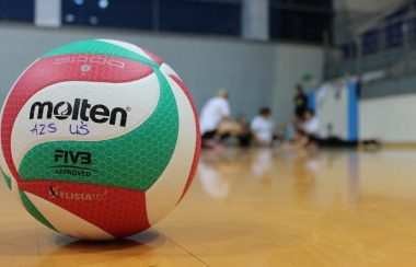 Ballon de volley-ball sur le sol, gymnase, jeunes joueurs en arrière-plan.