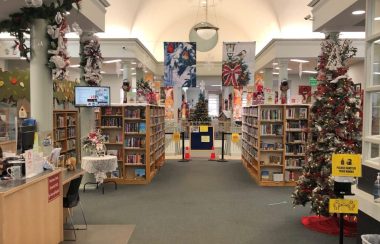 On aperçoit l'intérieur de la bibliothèque publique de Penetanguishene qui est décoré pour le temps des fêtes. On peut voit les rayons de livres et de films ainsi que le comptoir d'accueil qui se trouve sur la gauche.