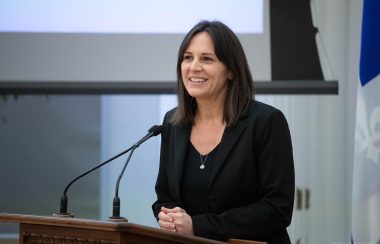Pictured is Isabelle Charest standing behind a podium speaking at a conference in Brome-Missisquoi. She is wearing a black t-shirt with a black blazer.