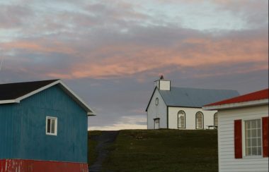 Une chapelle blanche et bleue entre deux bâtiments de bois sous un ciel rosée.