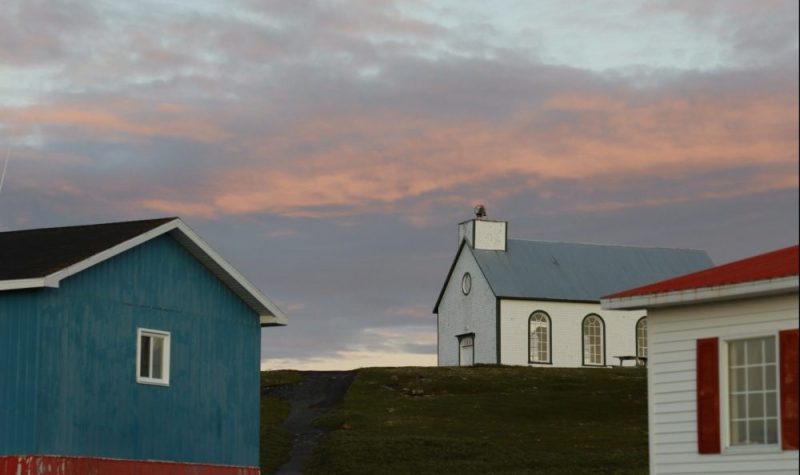 Une chapelle blanche et bleue entre deux bâtiments de bois sous un ciel rosée.
