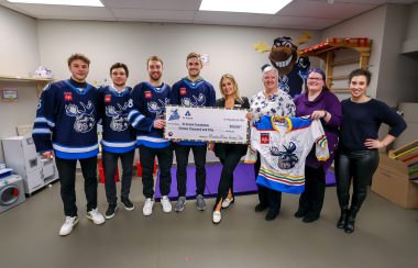 Les joueurs ont en main un chèque de 16 050,15 $ destiné à la Fondation St-Amant et portent le jersey des Manitoba Mooses, de couleur blanc et bleu. À côté d'eux, trois femmes sourient et tiennent le jersey de l'équipe de hockey.