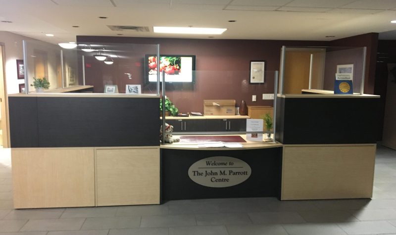 A black desk with wooden features in foreground with sign reading Welcome to The John M. Parrott Centre. Tile flooring and brown walls.