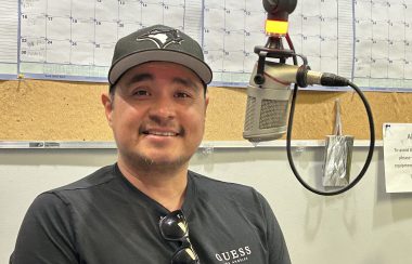 Man in dark grey top with grey baseball cap sits smiling toward the camera, a light beige background and a studio microphone is slightly to the right of him.