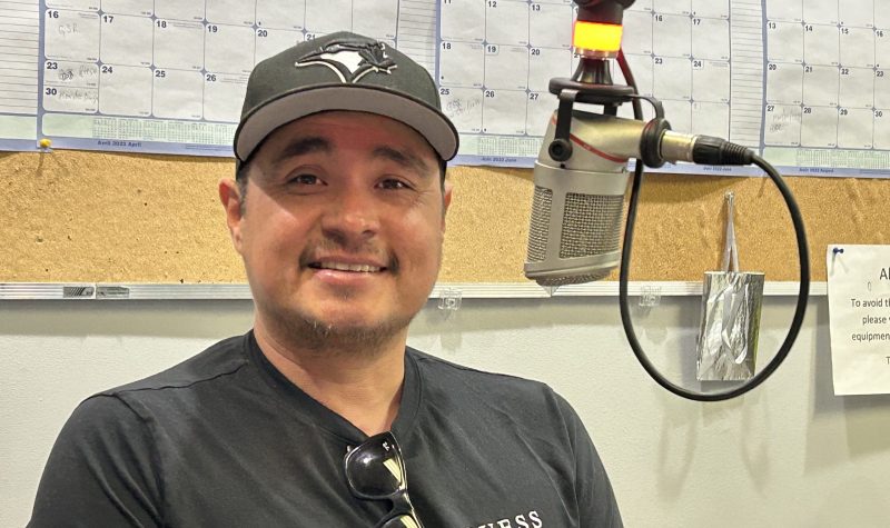 Man in dark grey top with grey baseball cap sits smiling toward the camera, a light beige background and a studio microphone is slightly to the right of him.