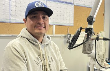 Man sitting at microphone in CKRZ FM station studio. He is wearing a white American Eagle sweatshirt and blue baseball cap.