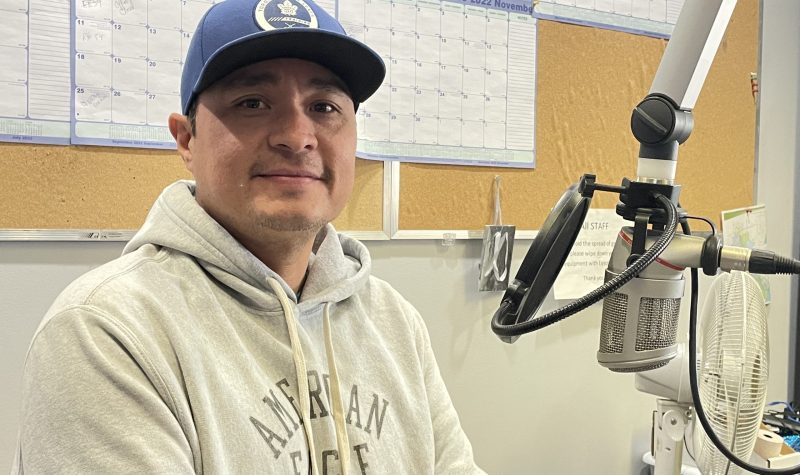 Man sitting at microphone in CKRZ FM station studio. He is wearing a white American Eagle sweatshirt and blue baseball cap.