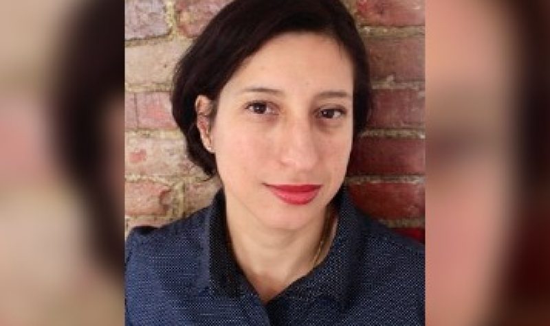 A portrait photo shows a woman with dark hair wearing a blue button-up top and standing against a red brick wall.