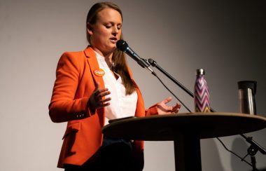Nelson-Creston MLA Brittny Anderson stands at a podium and a microphone on a stage against a grey and black background
