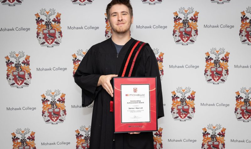 A man stands in front of backdrop holding graduation plaque.