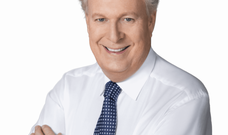 Jean Charest standing in front of a white background smiling with his arms crossed