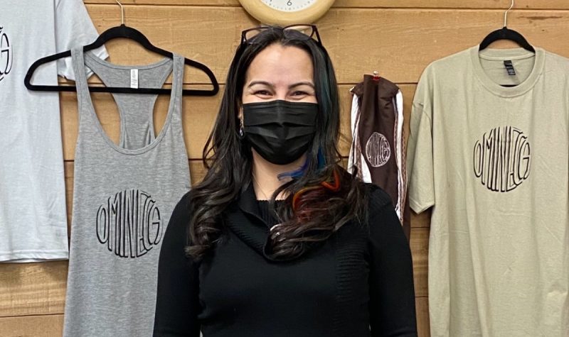 A person in a mask stands in front of printed t-shirt displayed for sale.