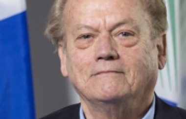 A professional headshot of Jim Gibson, standing in front of a Quebec flag and wearing a navy suit with a blue collared shirt.