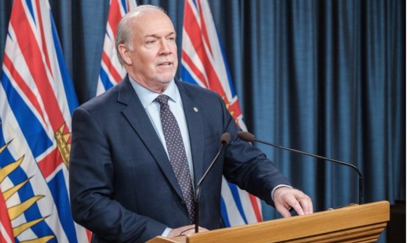 Premier John Horgan stands at a wooden podium during a press conference with BC flags and a blue curtain behind him