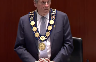 A person with a suit and large gold chain on. They are standing with a small desk in front. The background is dark brown.