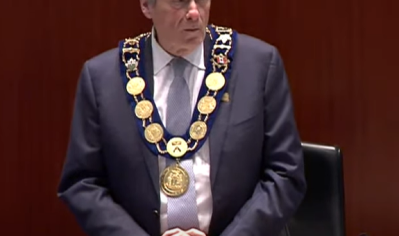 A person with a suit and large gold chain on. They are standing with a small desk in front. The background is dark brown.