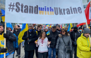 A group of people marching down a street wih a white banner being held by two people on each side. The banner reads out, Stand with Ukraine.