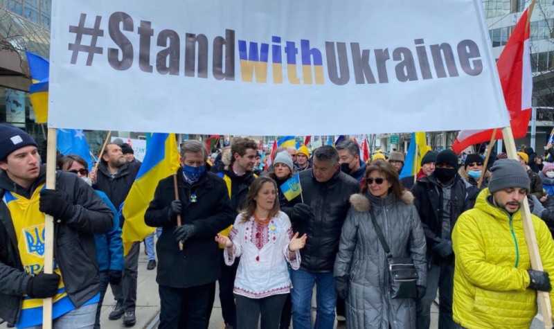 A group of people marching down a street wih a white banner being held by two people on each side. The banner reads out, Stand with Ukraine.