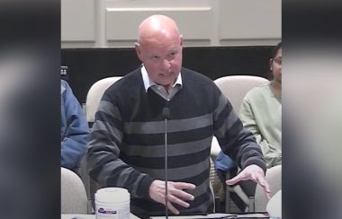 A bald man wearing a white collared shirt and V-neck sweater with dark coloured stripes gestures as he speaks in council chambers.