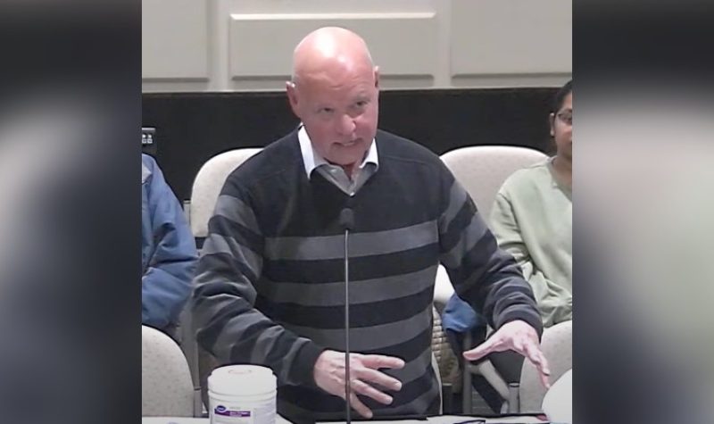 A bald man wearing a white collared shirt and V-neck sweater with dark coloured stripes gestures as he speaks in council chambers.