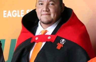 Lheidli T'enneh Councillor Joshua Seymour stands in front of orange step & shoot backdrop. He wears a black suit with white shirt and orange tie. Over it, is draped a traditional robe.