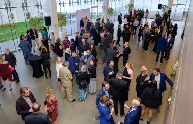 many people gathered, chatting in groups wearing formal clothing. They are in a large room surrounded by windows.