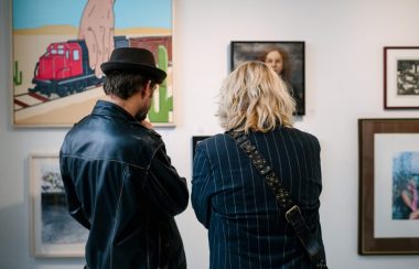 Two people with their backs to the camera observe paintings hung on a white wall. They are dressed in black and one is wearing a black hat.