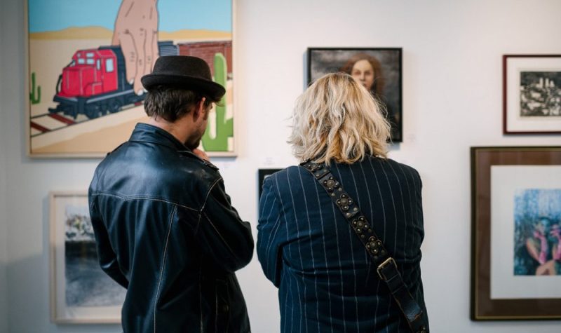 Two people with their backs to the camera observe paintings hung on a white wall. They are dressed in black and one is wearing a black hat.