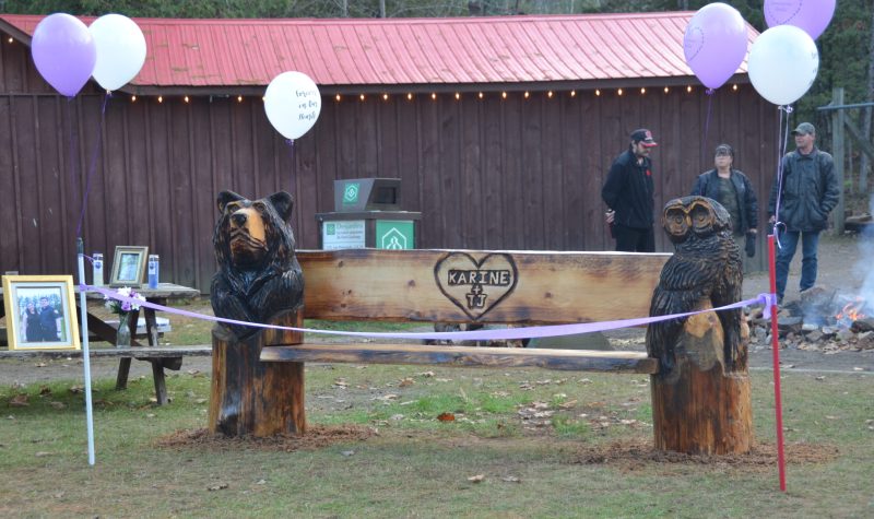 A photo of a rustic wooden bench with a carved bear on one end and an owl on the other with purple and white balloons.