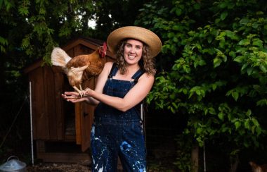 Karry Sawatsky holds a rooster in her hands in her community garden with trees behind her