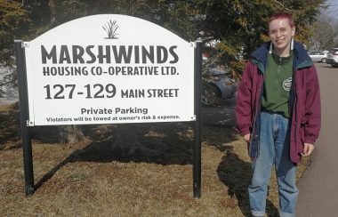 A woman stands next to a white and black sign for Marshwinds on a sunny day.