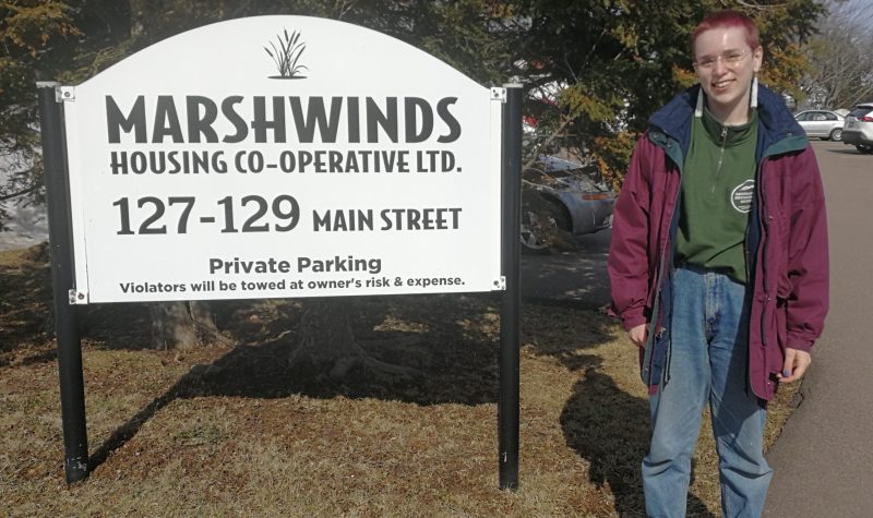 A woman stands next to a white and black sign for Marshwinds on a sunny day.