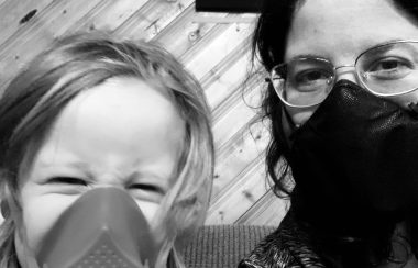 A young child wearing a respirator sits with her mother, who wears a face mask.