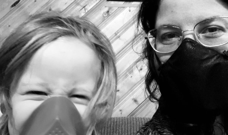 A young child wearing a respirator sits with her mother, who wears a face mask.