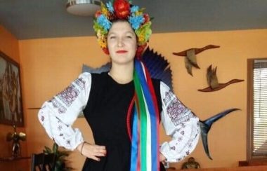 A full body shot of Katrina Soroka in traditional Ukrainian dress, with red boots, a black dress with white embroidered sleeves and a crown of flowers with multi-coloured ribbons attached at the back.