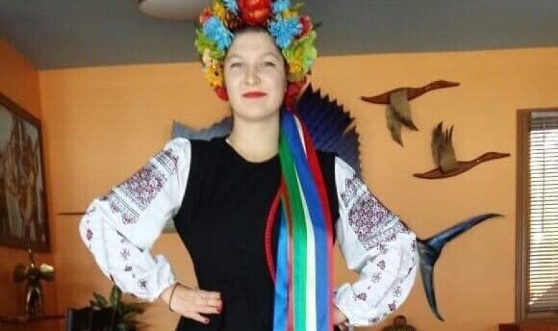 A full body shot of Katrina Soroka in traditional Ukrainian dress, with red boots, a black dress with white embroidered sleeves and a crown of flowers with multi-coloured ribbons attached at the back.