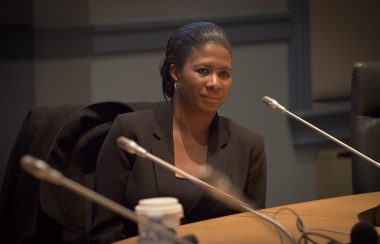Femme noire assise a une table on voit trois microphones devant elle, la table doit être celle du conseil municipal. La femme en question est Ketcia Peters